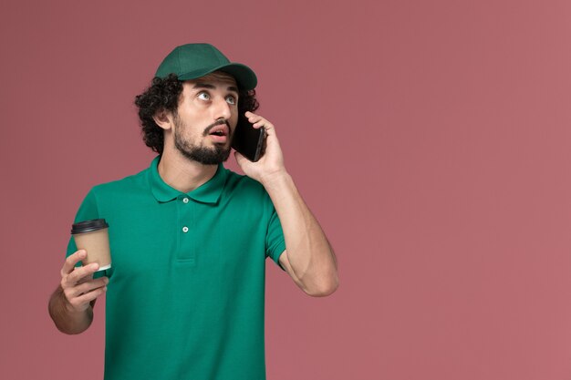 Front view male courier in green uniform and cape holding coffee cup and talking on the phone on pink background uniform delivery service