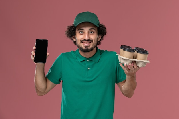 Front view male courier in green uniform and cape holding coffee cups with phone on the light-pink background service uniform delivery job