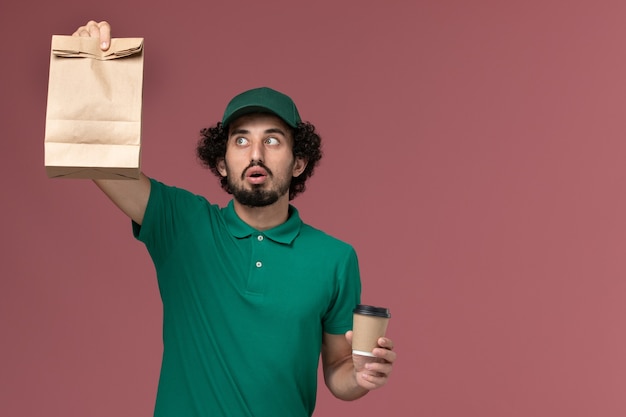 Front view male courier in green uniform and cape holding delivery coffee cup and food package on the light pink background uniform delivery service company job