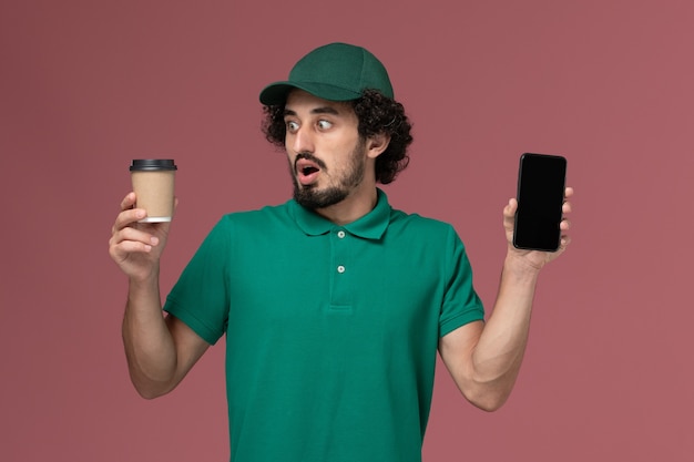 Front view male courier in green uniform and cape holding delivery coffee cup with phone on pink background uniform delivery job service