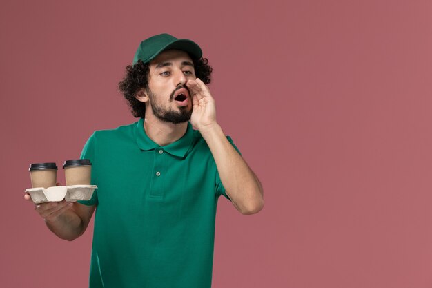 Front view male courier in green uniform and cape holding delivery coffee cups on the pink background service job male uniform delivery