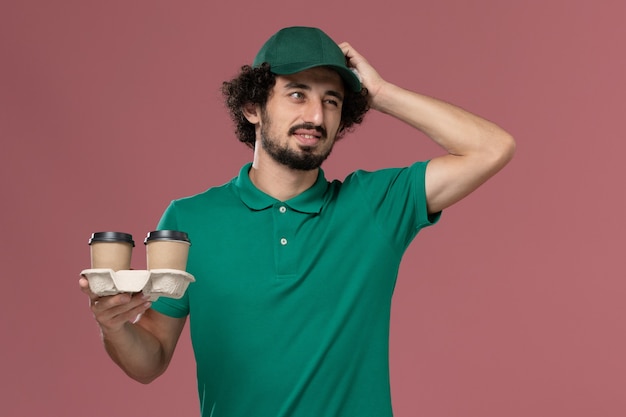 Front view male courier in green uniform and cape holding delivery coffee cups and thinking on pink background service job male worker uniform delivery