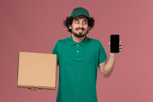 Front view male courier in green uniform and cape holding delivery food box and phone on pink background service worker uniform delivery