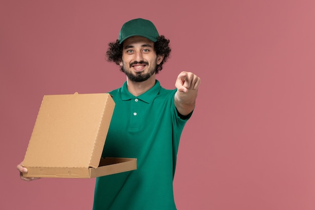 Front view male courier in green uniform and cape holding delivery food box on the pink desk service worker uniform delivery job