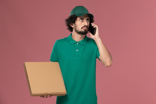 Free photo front view male courier in green uniform and cape holding delivery food box and talking on the phone on the pink floor service worker uniform delivery job