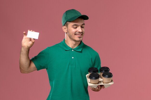 Front view male courier in green uniform holding brown coffee cups and card on the pink background  