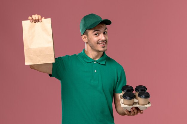 Front view male courier in green uniform holding brown coffee cups and food package smiling on pink background  