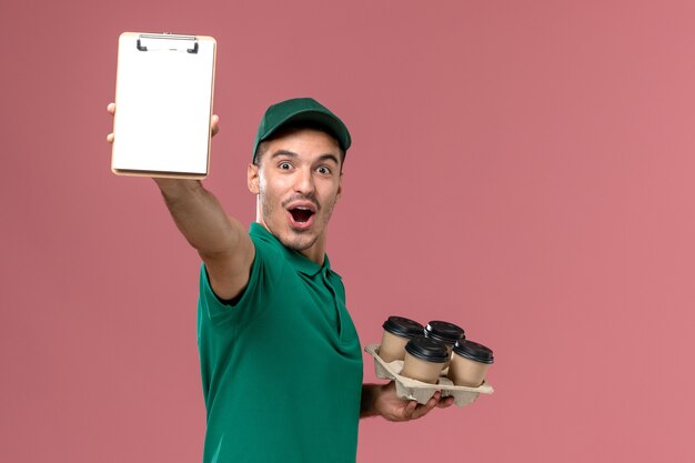 Front view male courier in green uniform holding brown coffee cups notepad on pink background   