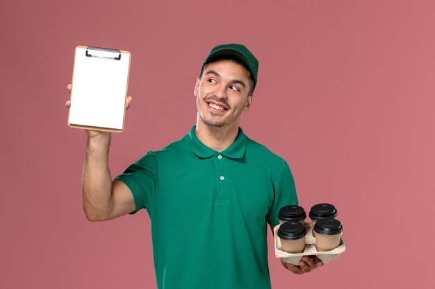 Front view male courier in green uniform holding brown coffee cups and notepad with a smile on pink background