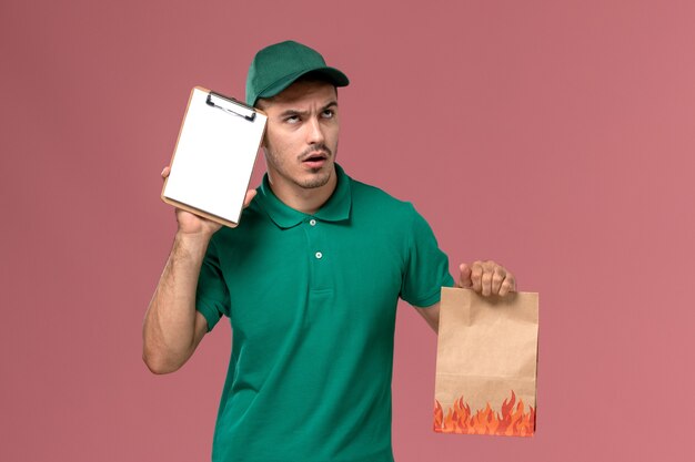 Front view male courier in green uniform holding food package and notepad thinking on light-pink background