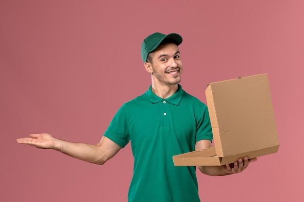 Front view male courier in green uniform holding and opening food box on light pink background   