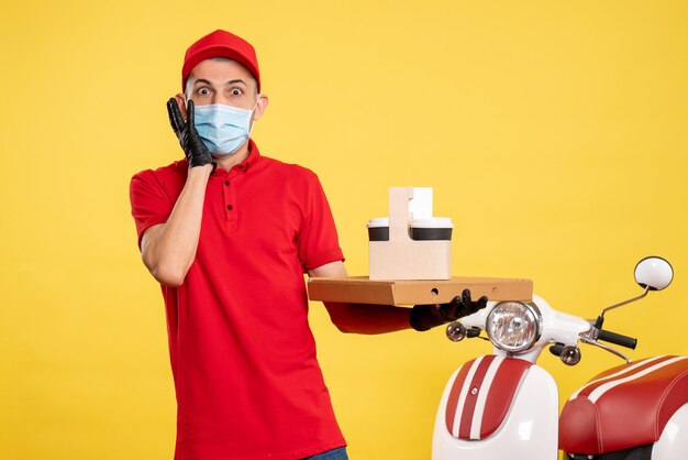 Front view male courier in mask with delivery coffee and box on yellow service covid- uniform color virus job