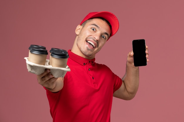 Free photo front view male courier in red uniform and cape holding delivery coffee cups with phone on the pink wall