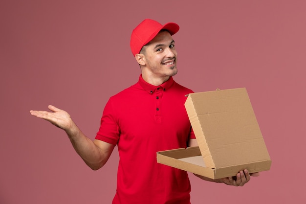 Front view male courier in red uniform and cape holding food box with smile on light-pink wall worker