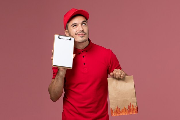 Front view male courier in red uniform and cape holding food package and notepad thinking on the pink wall 