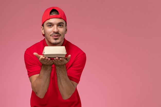 Free photo front view male courier in red uniform and cape holding little delivery package on the pink background.