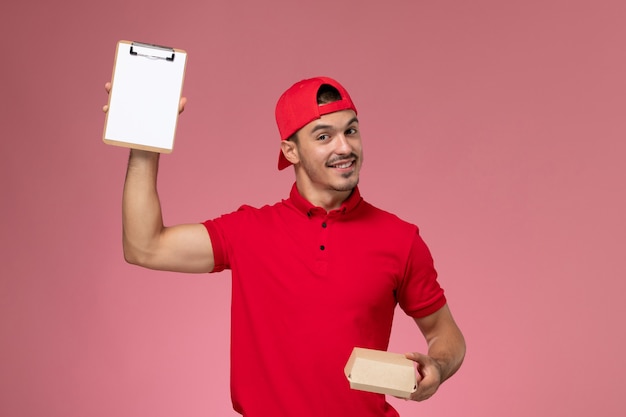 Front view male courier in red uniform and cape holding little delivery package with notepad on pink background.