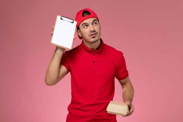 Front view male courier in red uniform and cape holding little delivery package with notepad thinking on pink background.