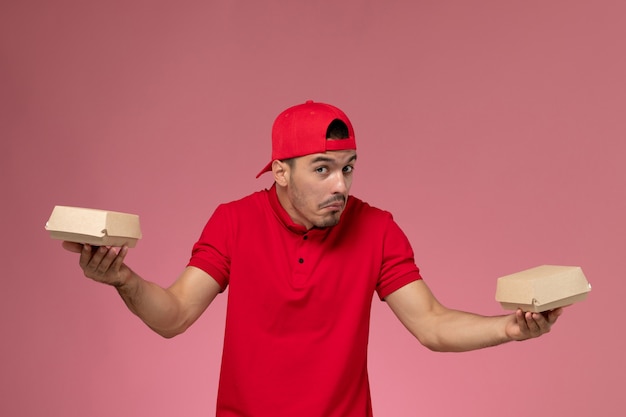 Free photo front view male courier in red uniform and cape holding little delivery packages on the pink background.