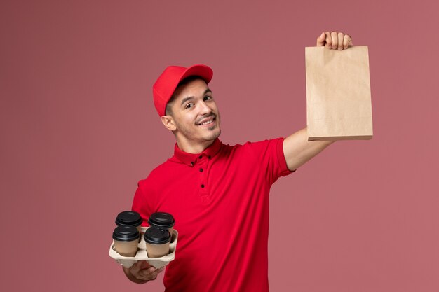 Front view male courier in red uniform holding brown delivery coffee cups with food package on pink desk service delivery job worker uniform