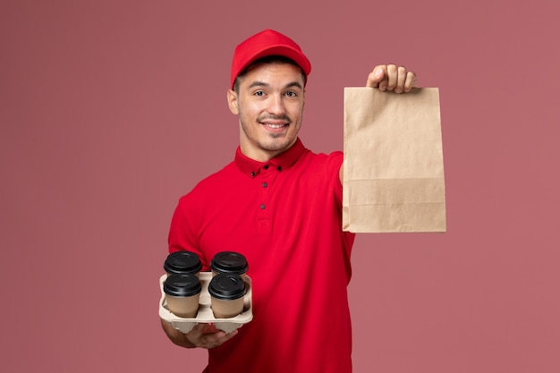 Front view male courier in red uniform holding brown delivery coffee cups with food package on the pink wall service delivery job worker uniform