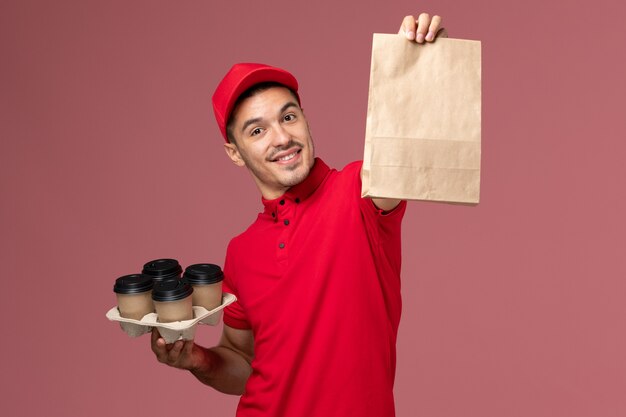 Front view male courier in red uniform holding brown delivery coffee cups with food package on the pink wall