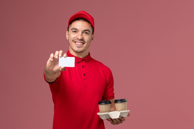 Front view male courier in red uniform holding brown delivery coffee cups with white card on light-pink wall 