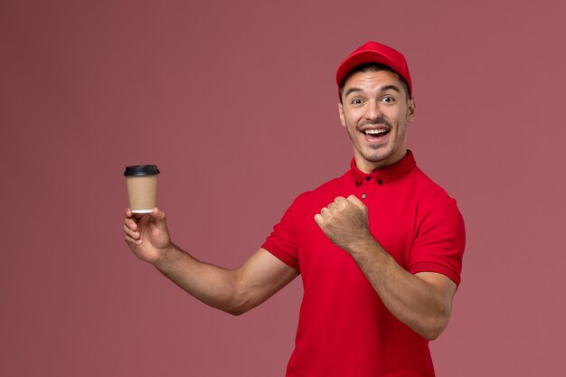 Front view male courier in red uniform holding delivery coffee cup and rejoicing on pink wall male worker