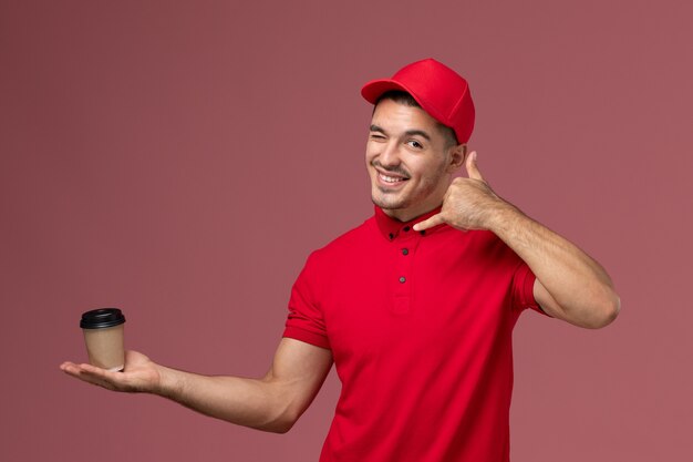 Front view male courier in red uniform holding delivery coffee cup winking on pink wall male job