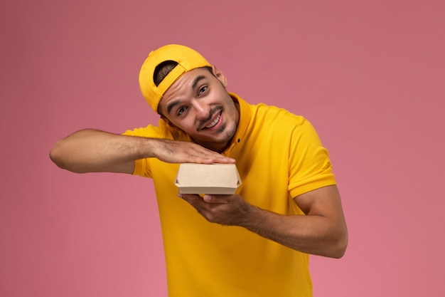 Free photo front view male courier in yellow uniform and cape holding little delivery food package on light-pink background.