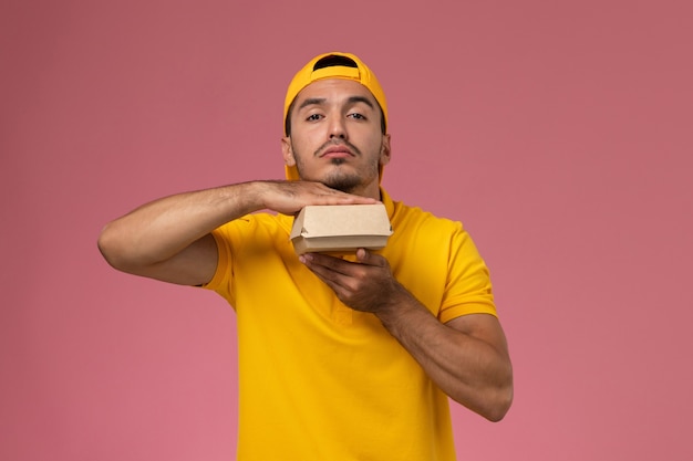 Free photo front view male courier in yellow uniform and cape holding little delivery food package on light-pink background.