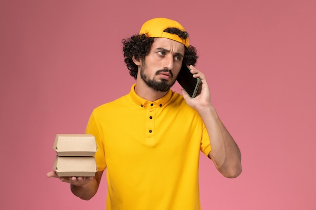 Front view male courier in yellow uniform cape with food packages on his hands talking on the phone on light-pink background.