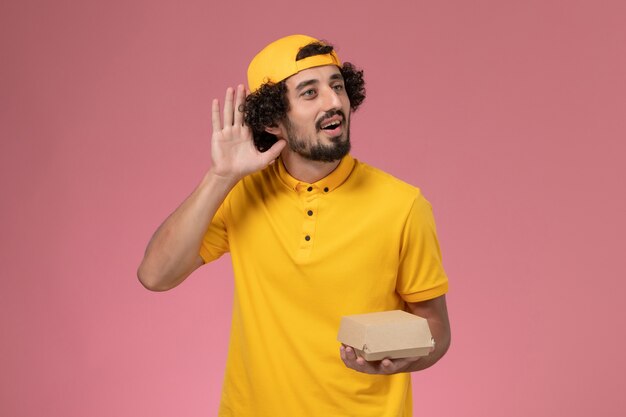 Front view male courier in yellow uniform and cape with little delivery food package on his hands on the pink background.