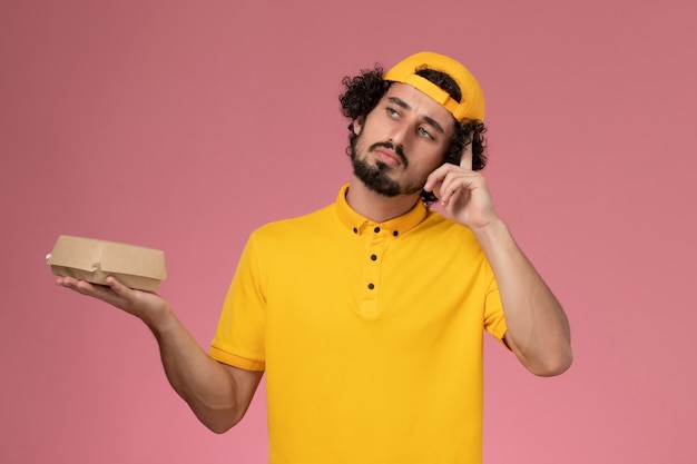 Front view male courier in yellow uniform and cape with little delivery food package on his hands thinking on the pink background.