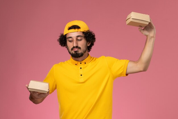 Free photo front view male courier in yellow uniform and cape with little delivery food packages on his hands on the pink background.