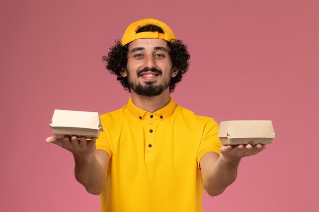 Free photo front view male courier in yellow uniform and cape with little delivery food packages on his hands on the pink background.