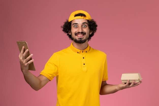 Free photo front view male courier in yellow uniform and cape with notepad and little delivery food package on his hands on pink background.