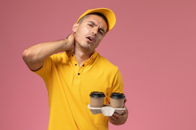 Front view male courier in yellow uniform holding delivery coffee cups having neckache on pink background 