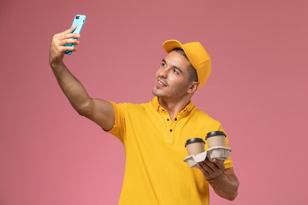 Front view male courier in yellow uniform holding delivery coffee cups taking a selfie on pink desk  