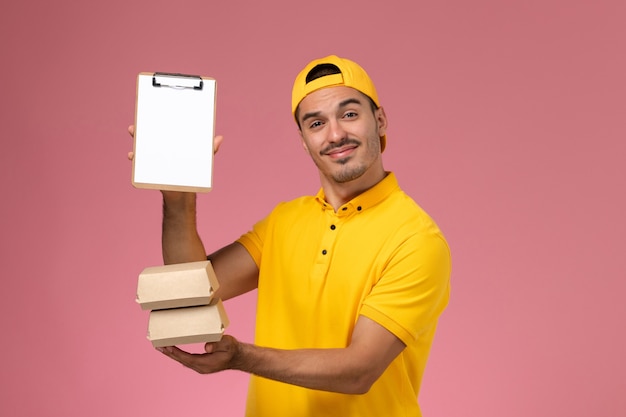 Front view male courier in yellow uniform holding little food packages and notepad on the light-pink background.