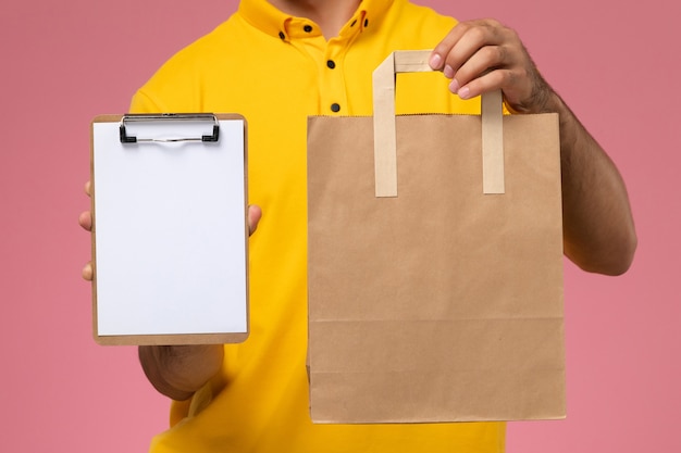 Front view male courier in yellow uniform holding little notepad and delivery food package on the pink background.