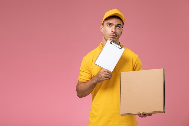 Front view male courier in yellow uniform thinking and holding notepad along with food delivery box on pink desk  