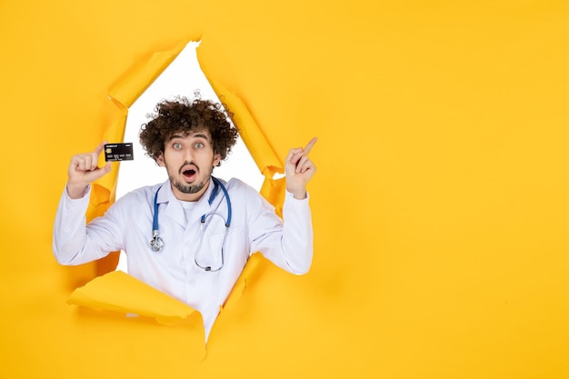 Front view male doctor in medical suit holding bank card on yellow ripped color medic health medicine virus disease