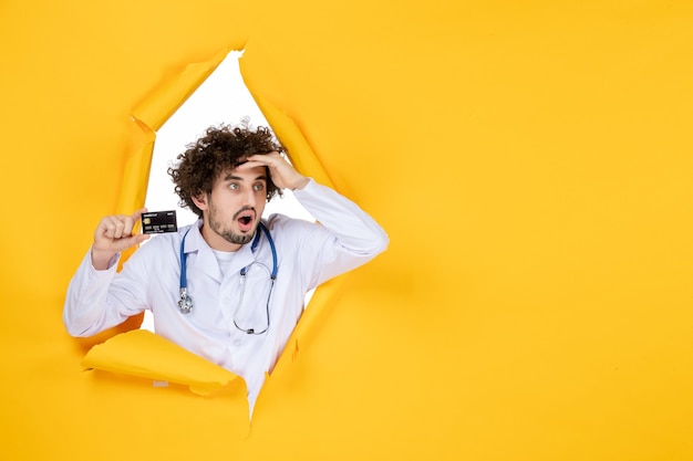 Front view male doctor in medical suit holding bank card on a yellow ripped color medic health medicine virus hospital disease