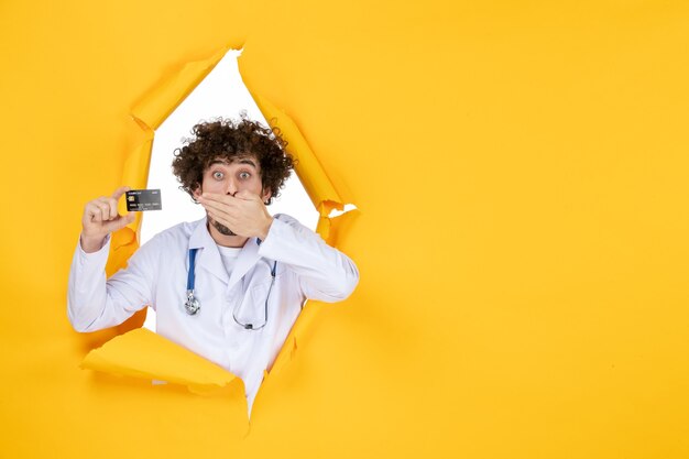 Front view male doctor in medical suit holding bank card on yellow ripped color medicine hospital disease health virus medic