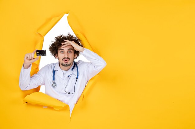 Front view male doctor in medical suit holding bank card on yellow ripped colors medic health medicine virus hospital disease