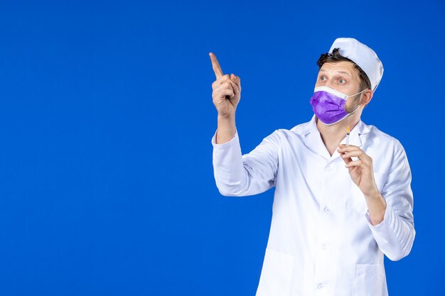 Front view of male doctor in medical suit and purple mask holding injection on blue 