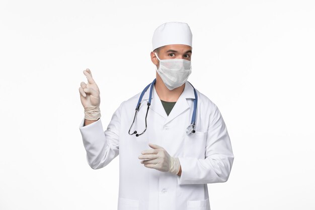 Front view male doctor in medical suit and wearing a mask against covid on light-white wall virus covid- illness disease pandemic