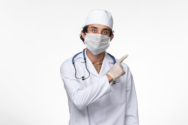 Front view male doctor in medical suit and wearing a mask as a protection from covid- on light white wall illness virus covid- pandemic