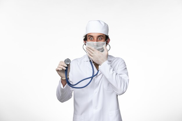 Front view male doctor in medical suit and wearing a mask as a protection from covid- using a stethoscope on the white desk virus illness covid- pandemic